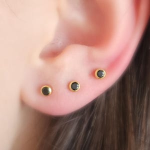 May include: Three small gold stud earrings with black gemstones. The earrings are worn in the earlobe and helix piercings.