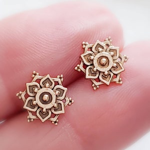 May include: A pair of gold-toned earrings with a detailed mandala design. Each earring features a circular mandala with intricate patterns and a small dot in the centre.