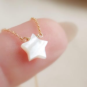 May include: A delicate gold chain necklace featuring a small, pearly white star pendant. The star has a five-point design and is made of a shimmering, iridescent material. The necklace is shown close-up, highlighting the fine details of the jewelry.