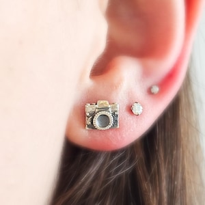 May include: A pair of silver earrings, one shaped like a camera and the other a small round stone.