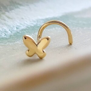 May include: A gold butterfly nose ring with a curved post. The ring is on a white background.
