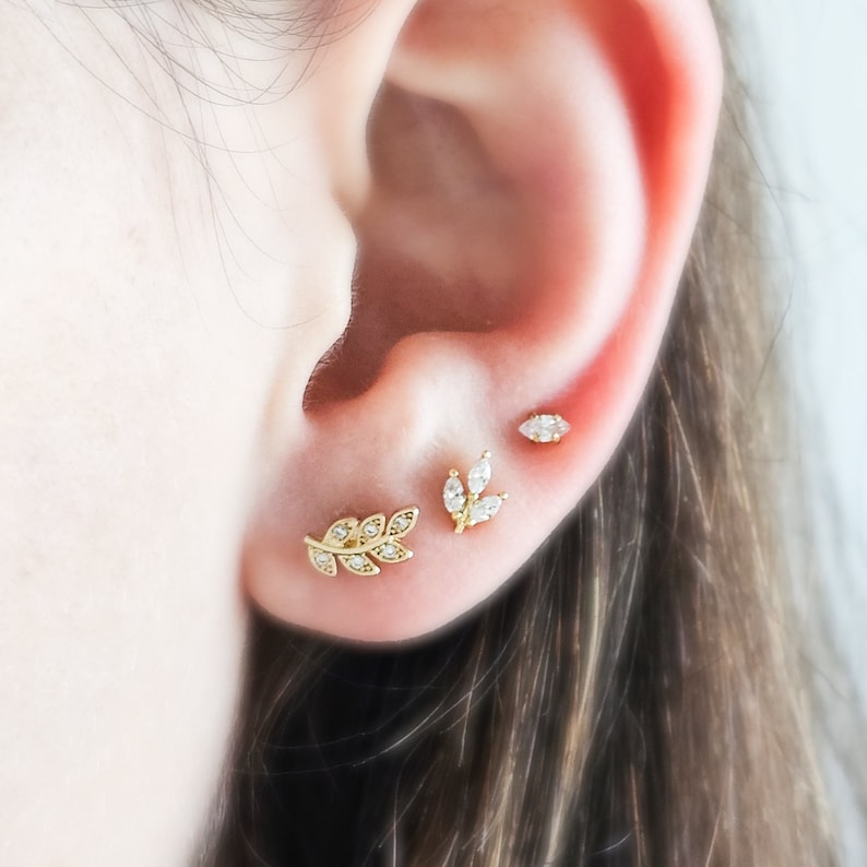 May include: Close-up of a person's ear with three gold earrings. The earrings are in the shape of leaves with small clear stones.