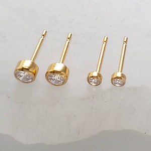May include: Four gold stud earrings with clear, round gemstones. The earrings are arranged in a row, with two larger and two smaller sizes. The studs have a simple, elegant design.