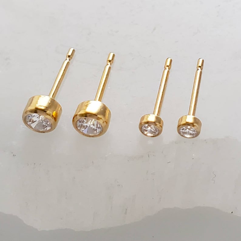 May include: Four gold stud earrings with clear, round gemstones. The earrings are arranged in a row, with two larger and two smaller sizes. The studs have a simple, elegant design.