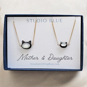 May include: A set of two gold necklaces with a black cat charm made of mother of pearl. The necklaces are in a blue box with the text "Studio Blue" and "Mother & Daughter ShopSomethingBlue.com".