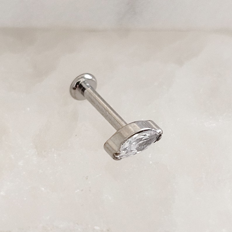 May include: A silver barbell with a clear marquise-shaped gemstone. The barbell is a body piercing jewelry item.