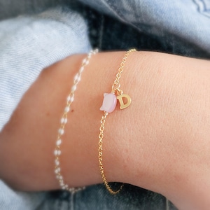 May include: Two delicate bracelets. One is a gold chain with a pink tulip charm and a gold letter "D" charm. The other is a white beaded chain. Both bracelets are on a wrist.