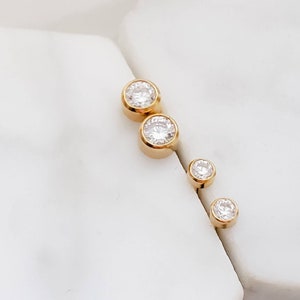 May include: Four gold-colored stud earrings with clear, round, sparkling gemstones. The earrings are arranged in a diagonal line, with two larger earrings at the top and two smaller ones below. The earrings are set against a white background.