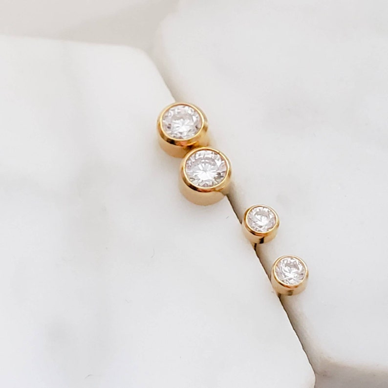 May include: Four gold-colored stud earrings with clear, round, sparkling gemstones. The earrings are arranged in a diagonal line, with two larger earrings at the top and two smaller ones below. The earrings are set against a white background.
