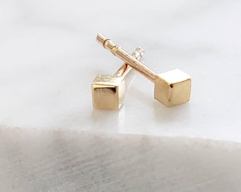 May include: Two gold cube stud earrings. Each earring features a small, square cube at the end of a thin post. The earrings are displayed on a white marble surface, with the light reflecting off the gold.