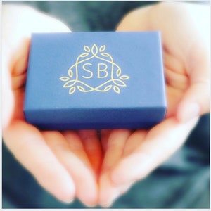May include: A small, dark blue box with a gold logo featuring the letters "SB" surrounded by stylized leaves. The box is being held in the palms of two hands.