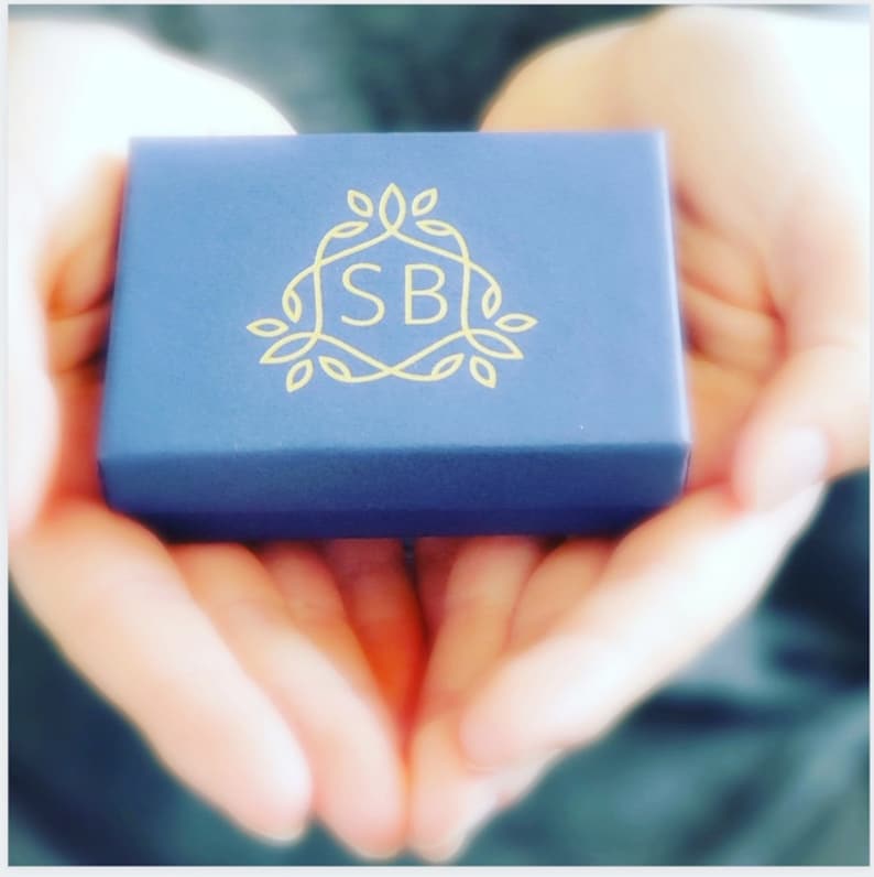 May include: A small, dark blue box with a gold logo featuring the letters "SB" surrounded by stylized leaves. The box is being held in the palms of two hands.