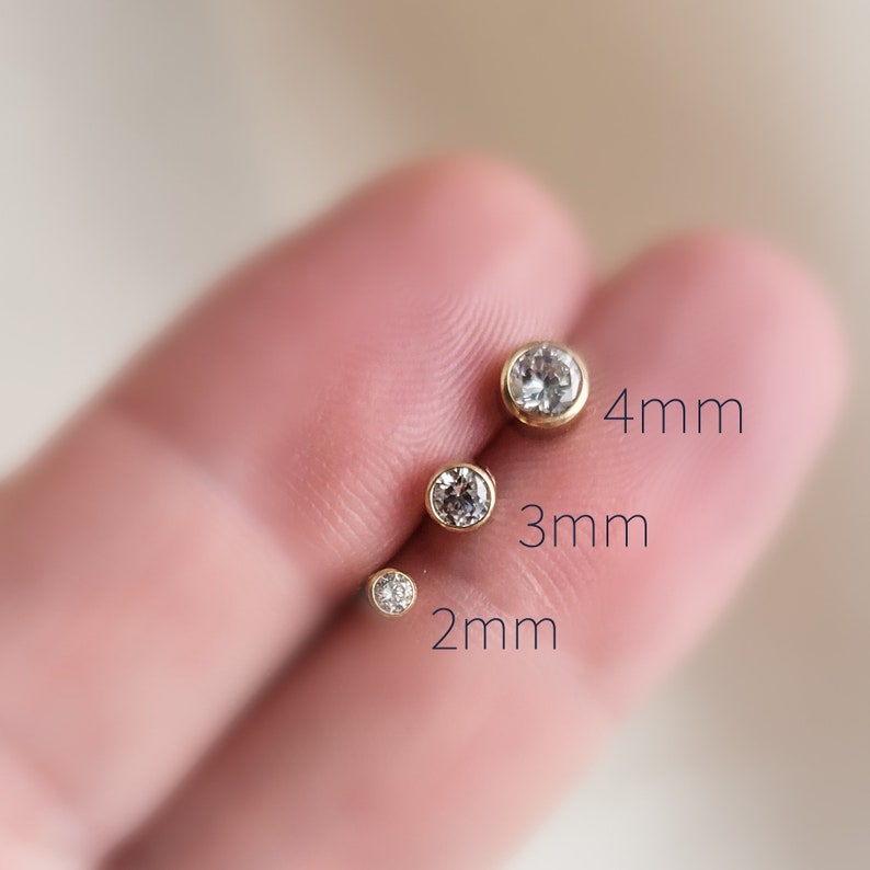 May include: Four gold-colored stud earrings with clear, round gemstones. The earrings are displayed in sizes 2mm, 3mm, and 4mm. The earrings are set in a gold bezel setting.