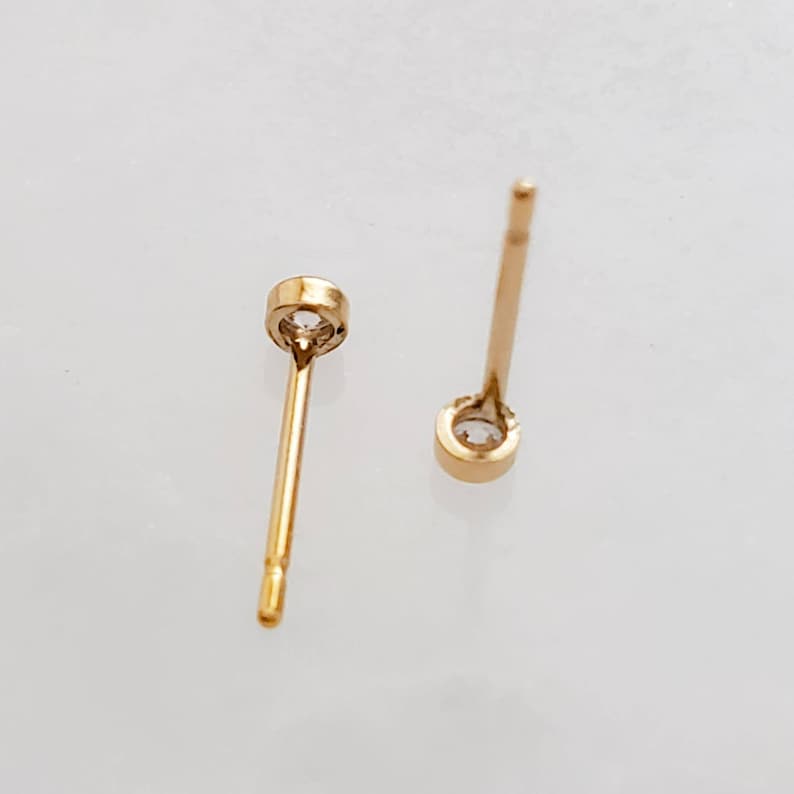May include: A pair of gold-colored stud earrings with a small, clear gemstone set in a bezel. The earrings have a simple, elegant design, with a post for insertion into the ear. The background is a light, neutral color.