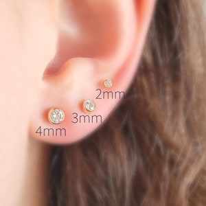 May include: Close-up of an ear with three gold stud earrings, each with a clear, round gemstone. The earrings are 2mm, 3mm, and 4mm in size. The ear is in focus, with the background blurred.