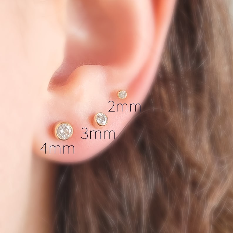 May include: Close-up of an ear with three gold stud earrings, each with a clear, round gemstone. The earrings are 2mm, 3mm, and 4mm in size. The ear is in focus, with the background blurred.
