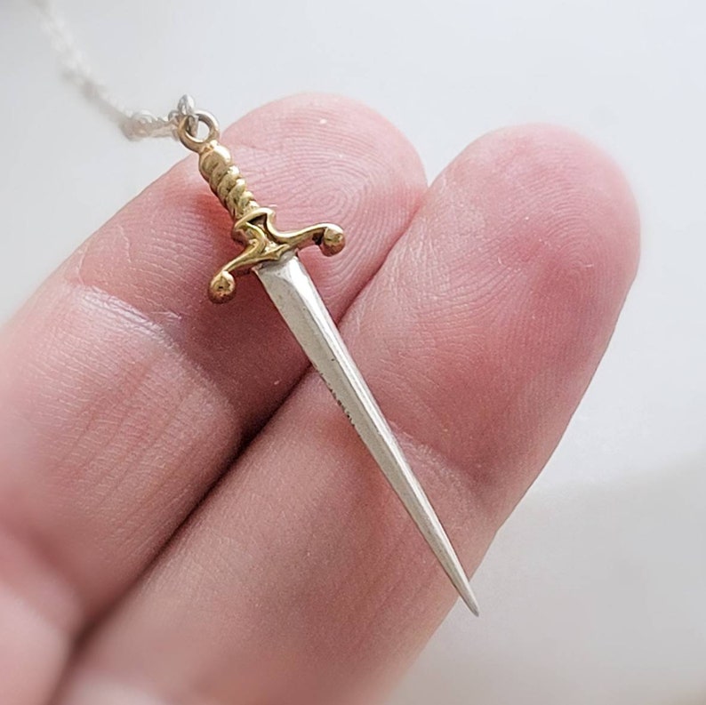 May include: A silver and gold sword pendant necklace. The sword has a detailed hilt and a pointed tip.