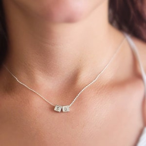 May include: A delicate silver necklace featuring two small square charms with the letters "S" and "B". The necklace is worn around the neck, showcasing the personalized pendant. The chain is thin and the pendant is centered.