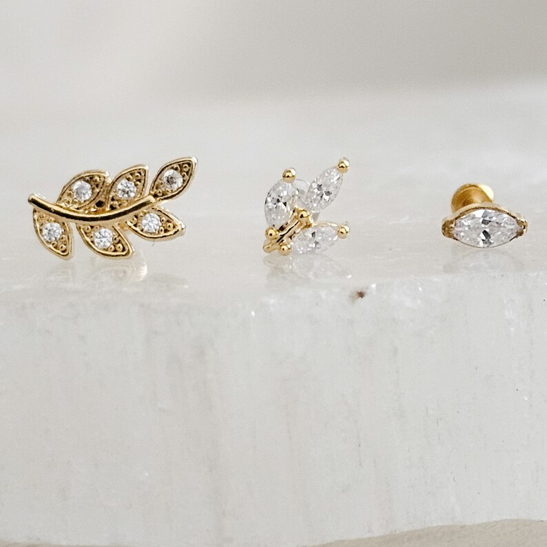 May include: Three gold-toned earrings with clear crystal accents. The earrings feature a leaf design, a cluster of crystals, and a single crystal.