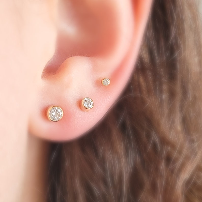 May include: Close-up of three gold stud earrings with clear, round, sparkling gemstones. The earrings are set in a bezel setting and are of varying sizes, creating a graduated look on the ear.