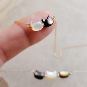 May include: A delicate gold necklace featuring two cat-shaped pendants. One pendant is white, the other is black. The necklace is displayed on a finger, with a blurred image of a similar necklace in the background.