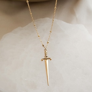 May include: A gold necklace with a sword pendant. The necklace is made of small, round beads and the sword is detailed with a twisted handle and a pointed tip.