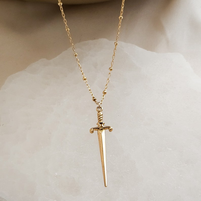 May include: A gold necklace with a sword pendant. The necklace is made of small, round beads and the sword is detailed with a twisted handle and a pointed tip.