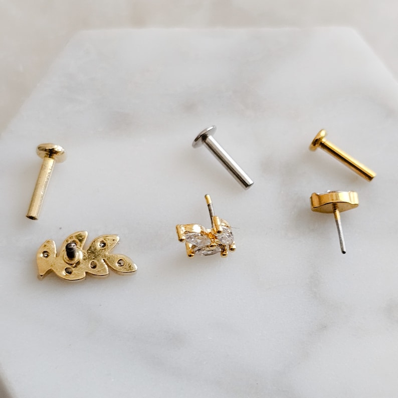 May include: Four gold and silver body jewelry pieces, including a leaf design, a crystal design, and two straight posts.