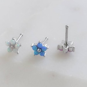 May include: Three silver nose rings with flower designs. The flowers are made of opal stones. The first flower is white, the second is blue, and the third is pink.
