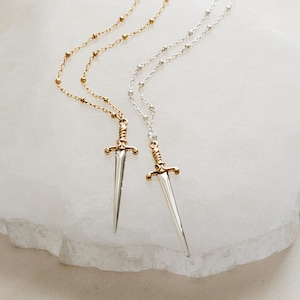 May include: Two delicate necklaces with a sword pendant. One necklace is gold and the other is silver. Both necklaces feature a small, detailed sword charm.