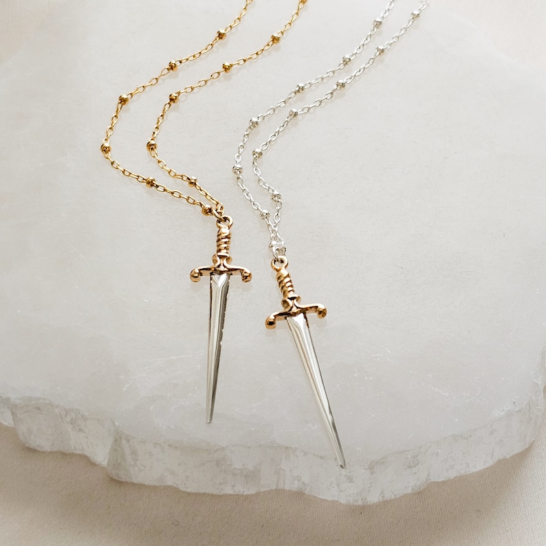 May include: Two delicate necklaces with a sword pendant. One necklace is gold and the other is silver. Both necklaces feature a small, detailed sword charm.