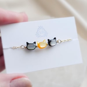 May include: A delicate gold-tone chain bracelet featuring three small cat-shaped beads. The outer two beads are dark grey with a pearlescent finish, while the centre bead is a vibrant orange. The bracelet is displayed on a white card with an SB logo.
