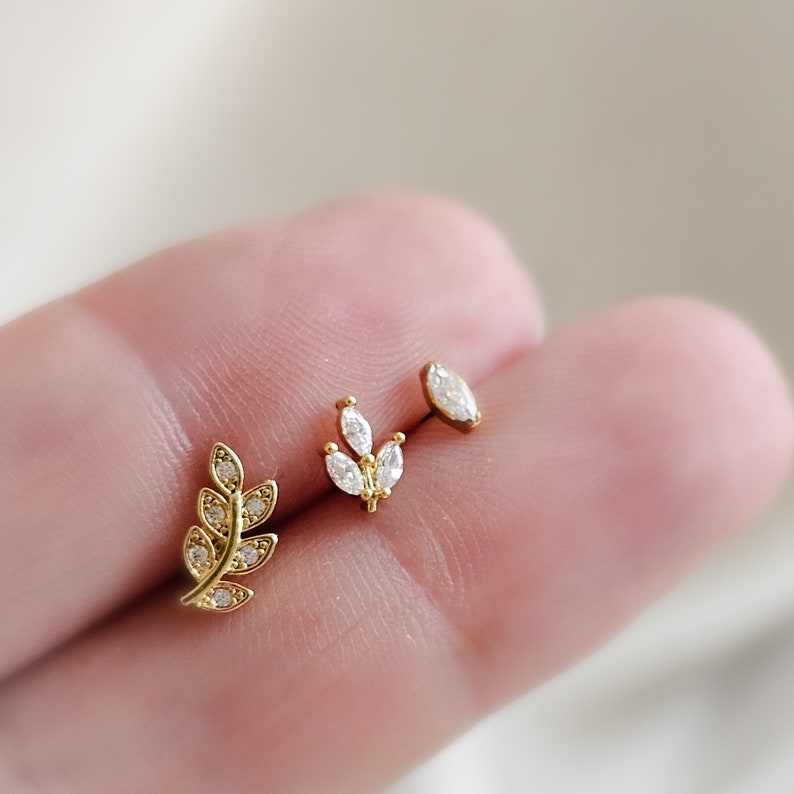 May include: Three gold earrings with clear crystal accents. The earrings feature a small leaf design, a larger leaf design, and a single oval-shaped crystal.