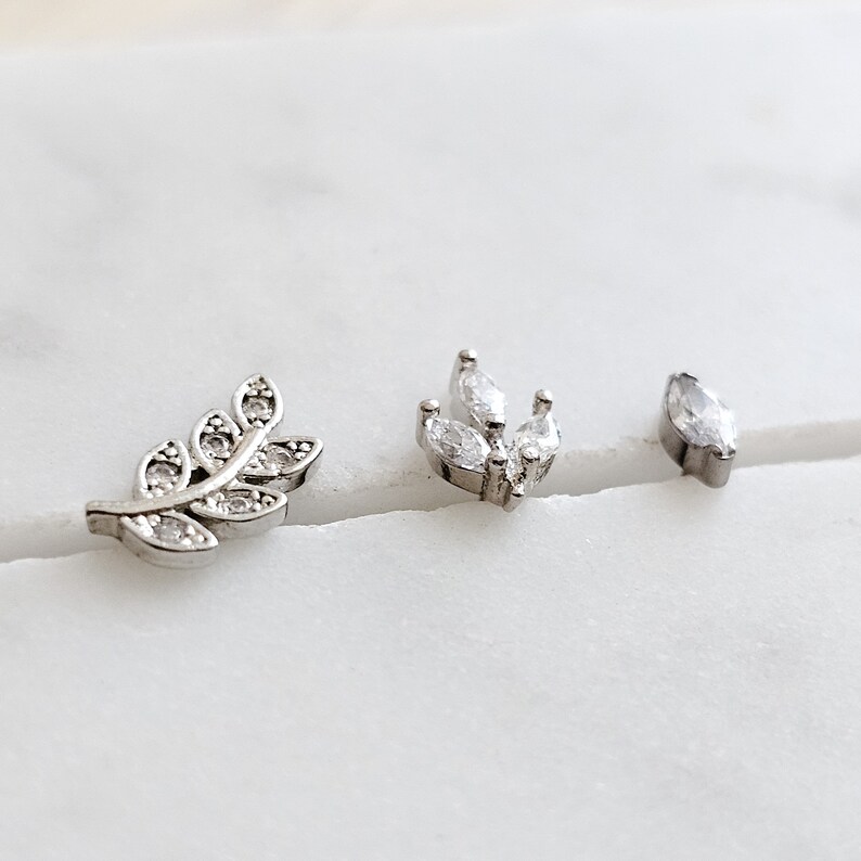 May include: Three silver body jewelry pieces with clear crystal accents. The pieces are a leaf, a cluster of leaves, and a single marquise-shaped stone.