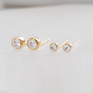 May include: Two pairs of gold stud earrings with clear, round gemstones. The earrings are set in a bezel setting, with one pair slightly larger than the other. The studs are displayed on a white surface.