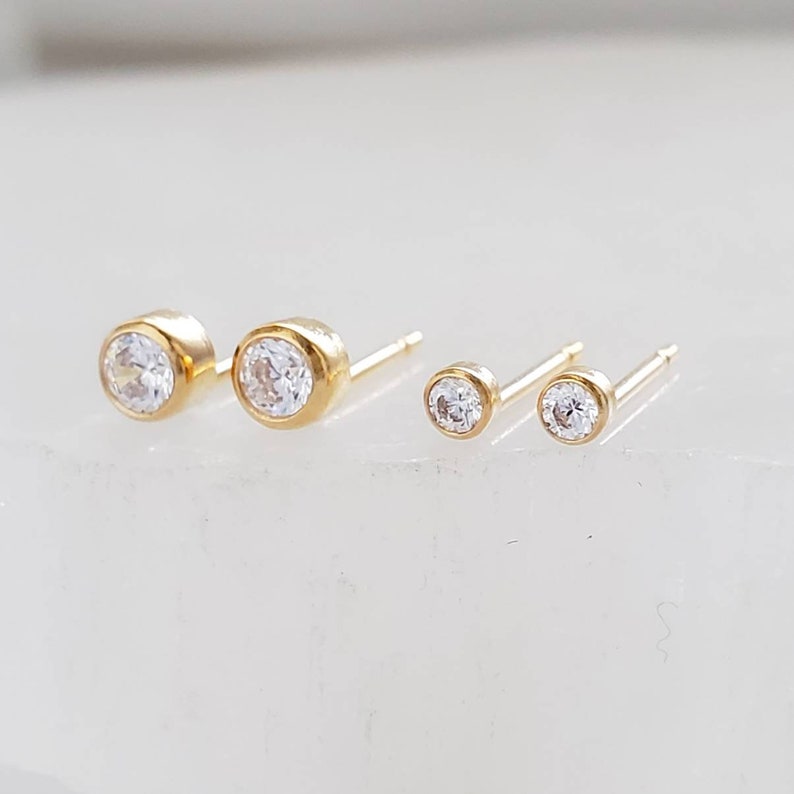 May include: Two pairs of gold stud earrings with clear, round gemstones. The earrings are set in a bezel setting, with one pair slightly larger than the other. The studs are displayed on a white surface.
