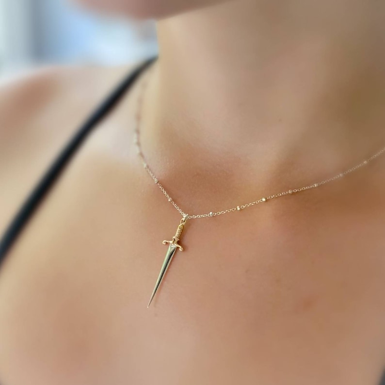May include: A gold sword pendant necklace on a silver chain. The necklace is worn by a person with a black tank top.