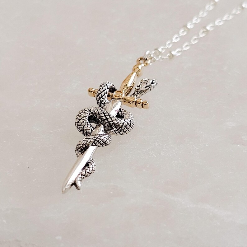 May include: A silver-toned pendant necklace featuring a detailed snake coiled around a sword. The sword's hilt is gold-toned. The snake's scales are intricately detailed. The necklace chain is silver. The pendant is displayed on a white surface.