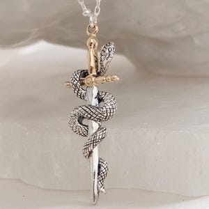 May include: A silver-toned pendant necklace featuring a detailed snake coiled around a sword. The sword's hilt and the snake's head are gold-colored. The necklace hangs from a delicate silver chain, creating a striking, symbolic accessory.