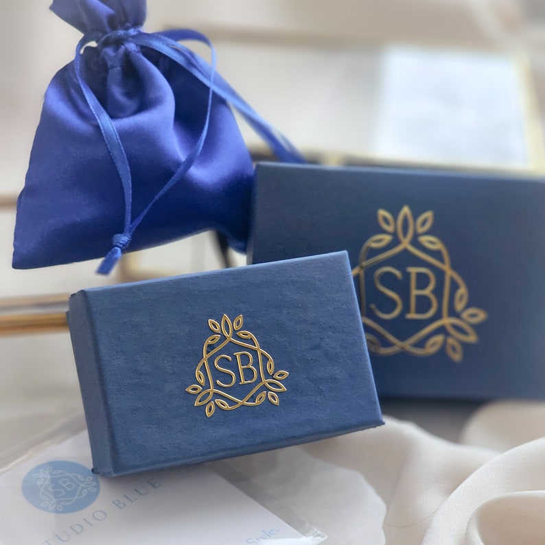 May include: Two blue boxes with gold foil lettering. The boxes have a gold logo with the letters "SB" inside a stylized design. The logo is surrounded by leaves. The boxes are on a white surface.