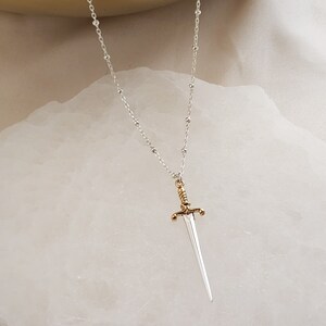 May include: A silver chain necklace with a gold-tipped sword pendant. The sword is silver with a gold handle and a pointed tip.