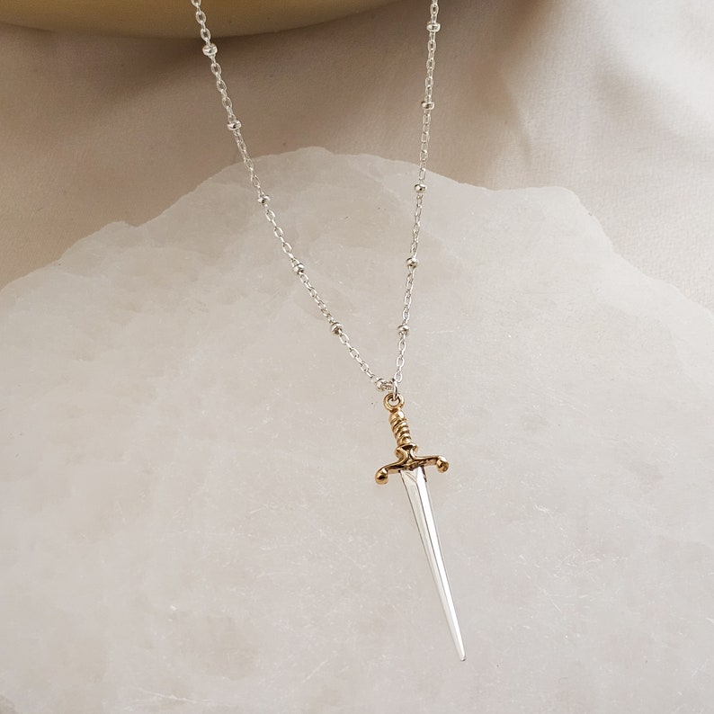 May include: A silver chain necklace with a gold-tipped sword pendant. The sword is silver with a gold handle and a pointed tip.