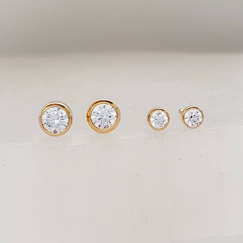 May include: Four gold-colored stud earrings with clear, round gemstones. The earrings vary in size, with the largest two having a bezel setting. The smaller earrings also have a bezel setting. The gemstones sparkle.