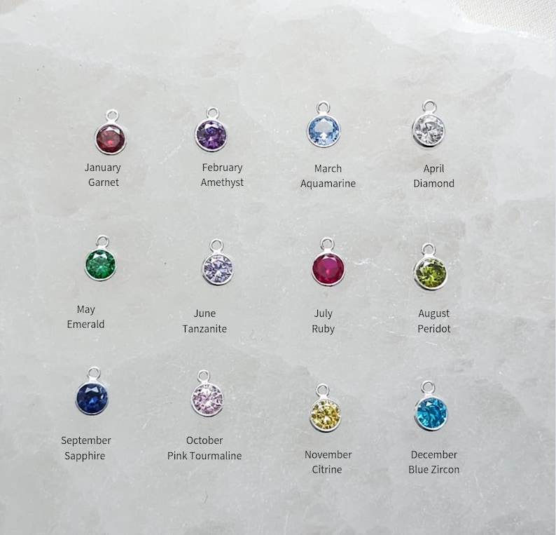 May include: A collection of twelve gemstone charms, each with a silver loop. The charms represent birthstones for each month, including Garnet, Amethyst, Aquamarine, Diamond, Emerald, Tanzanite, Ruby, Peridot, Sapphire, Pink Tourmaline, Citrine, and Blue Zircon.