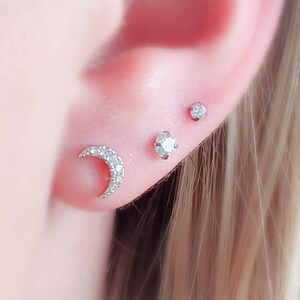 May include: Three silver earrings with clear cubic zirconia stones. The earrings are in the shape of a crescent moon, a small round stone, and a larger round stone.