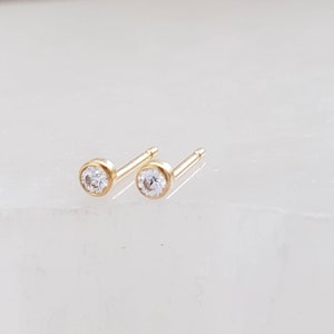 May include: A pair of small, gold-colored stud earrings. Each earring features a round, clear gemstone set in a bezel setting. The earrings are displayed on a light-colored surface.