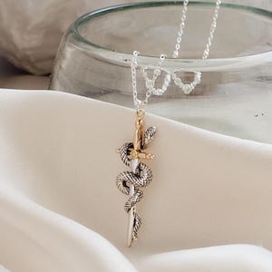 May include: A silver necklace with a pendant featuring a sword entwined by a snake. The sword has a gold-colored hilt and a silver blade. The snake is detailed with scales. The necklace is displayed on a white fabric.