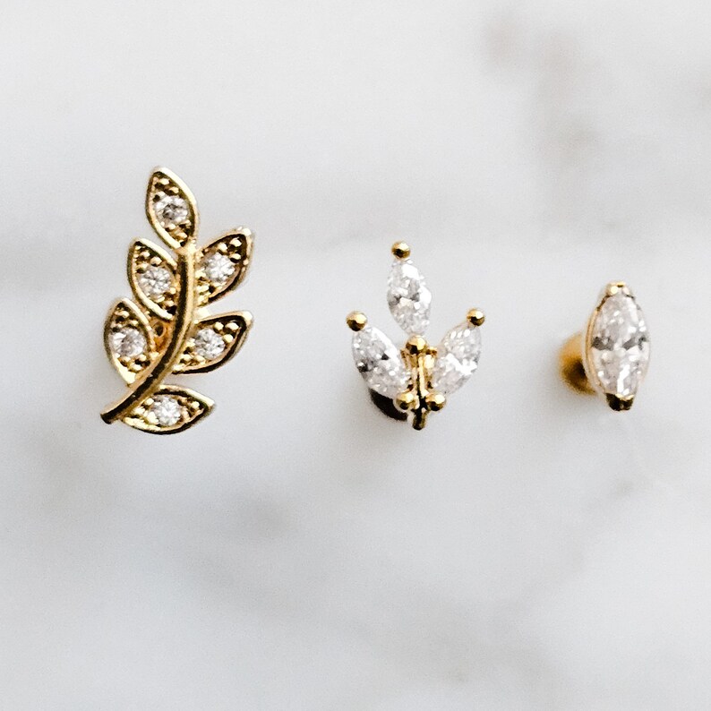 May include: Three gold-toned earrings with clear crystal accents. The earrings feature a leaf design, a flower design, and a single marquise-shaped crystal.