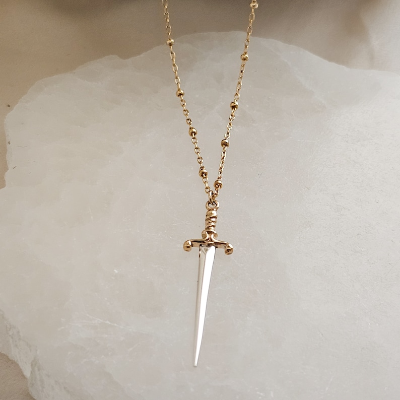 May include: A gold chain necklace with a silver sword pendant. The sword has a detailed handle and a pointed tip.