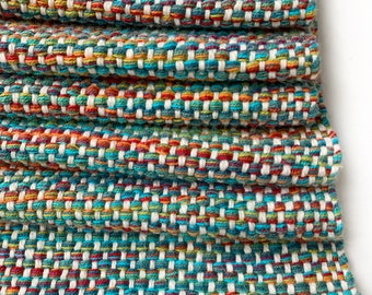 May include: A folded, handwoven textile with a vibrant, multicolored pattern. The design features a mix of teal, red, orange, yellow, and white threads, creating a textured, geometric appearance. The fabric is tightly woven, suggesting durability and a handcrafted aesthetic.